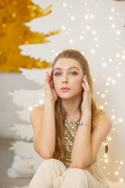 girl in a shiny evening dress. Christmas Eve. Cozy holiday at the fur-tree with lights and gold decor - Foto, Imagen