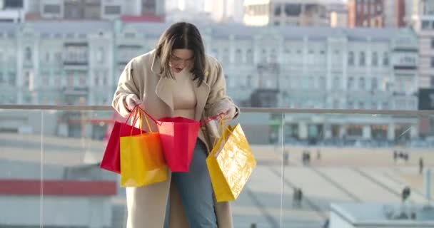 Satisfied Caucasian woman looking into colorful shopping bags and smiling. Young female shopaholic standing on city street with purchases. Shopaholism, lifestyle. Cinema 4k ProRes HQ. - Filmagem, Vídeo