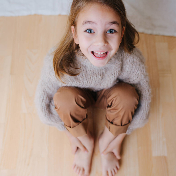 Grimacing little girl looks up while sitting on a parquet, holding her bare feet - Photo, Image