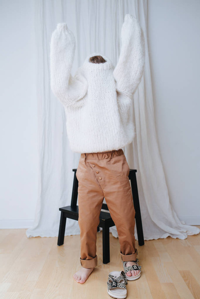 Funny little girl struggling to put on white fluffy knitted sweater. - Foto, Imagen