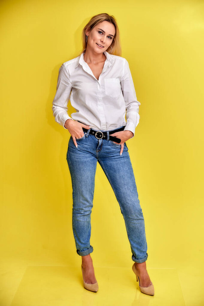 Full-length art portrait of a pretty smiling blonde woman in blue jeans and a white shirt stands on a yellow background. Right in front of the camera with emotions. - Фото, изображение