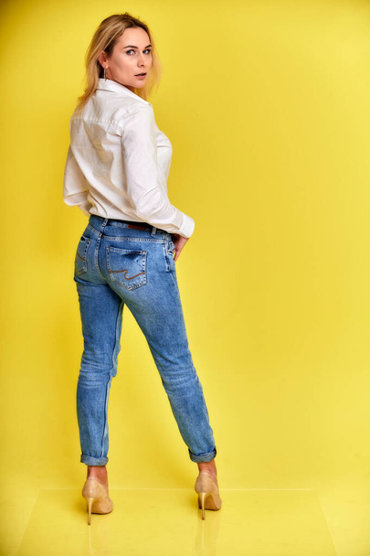 Full-length art portrait of a pretty smiling blonde woman in blue jeans and a white shirt stands on a yellow background. Right in front of the camera with emotions. - Photo, image