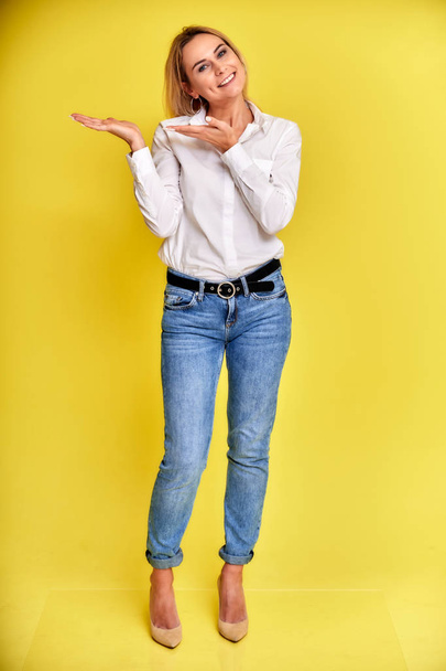 Full-length art portrait of a pretty smiling blonde woman in blue jeans and a white shirt stands on a yellow background. Right in front of the camera with emotions. - Photo, Image