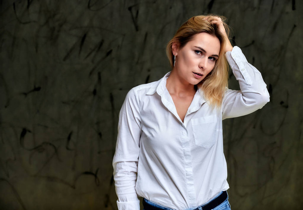 Portrait of a pretty smiling blonde woman in a white shirt with flying hair standing on a gray alternative background. Right in front of the camera with emotions. - Photo, Image