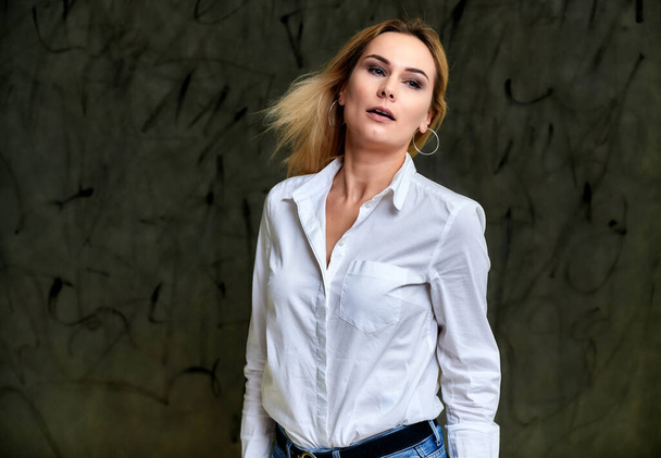 Portrait of a pretty smiling blonde woman in a white shirt with flying hair standing on a gray alternative background. Right in front of the camera with emotions. - Foto, Bild
