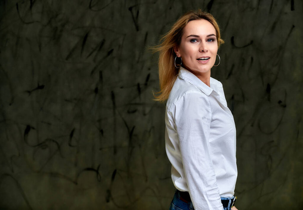 Portrait of a pretty smiling blonde woman in a white shirt with flying hair standing on a gray alternative background. Right in front of the camera with emotions. - Valokuva, kuva
