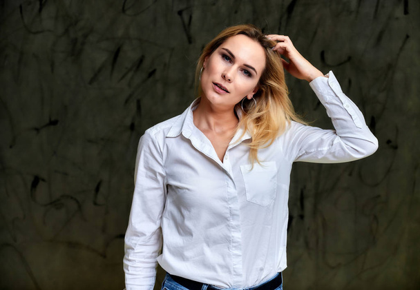 Portrait of a pretty smiling blonde woman in a white shirt with flying hair standing on a gray alternative background. Right in front of the camera with emotions. - Фото, изображение