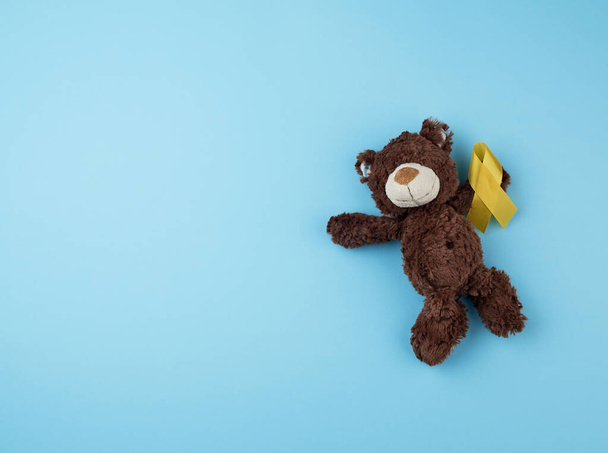 Το μικρό καφέ αρκουδάκι κρατά στην πατούσα του μια κίτρινη κορδέλα διπλωμένη  - Φωτογραφία, εικόνα