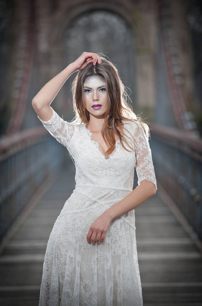Lovely young lady wearing elegant white dress enjoying the beams of celestial light and snowflakes falling on her face. Pretty brunette girl in long wedding dress posing on a bridge in winter scenery - Foto, imagen