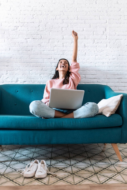 Pretty young woman celebrating something while using her laptop on the sofa at home. - Foto, imagen