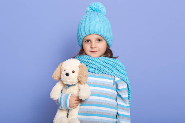 Studio shot of little girl with her soft dog toy in hands, standing against blue wall wearing striped shirt, scarf and cap with pom pom, looking directly at camera, has dark hair, plays with her toy. - Fotografie, Obrázek