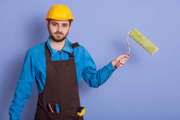 Studio shot of young unshaven builder dresses shirt, brown apron and yellow protective helmet, standing against blue wall and holding painting roller in hands,ready to paint walls. Building concept. - Photo, image