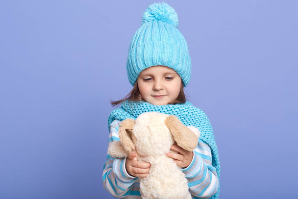 Horizontalshot of baby girl holding her soft dog toy in hands and looking at it, wearing warm clothing, isolated over blue studio background. Family, child, childhood and parenthood concept. - Valokuva, kuva
