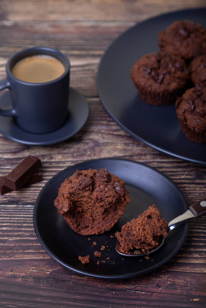 Chocolate muffins on a black plate. Homemade baking. In the background is a cup of coffee and a plate with muffins. Wooden background. Selective focus, close up. - Foto, Imagem