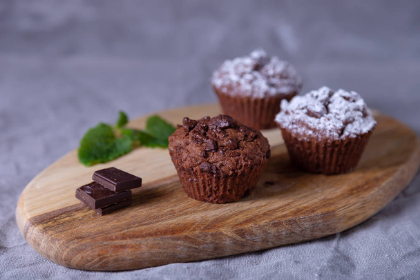 Chocolate muffins on a wooden board, strewed with icing sugar. Homemade baking. In the background is a cup of coffee and a plate with muffins. Wooden background. Selective focus, close up. - Foto, Imagem
