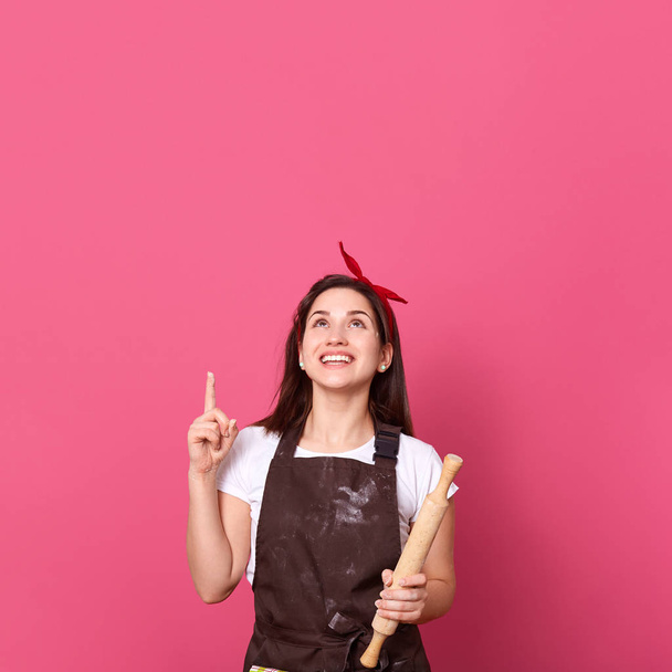Image of smiling female chef, cook or baker with rolling pin in hands pointing up with her index finger and looking up, isolated over pink background, copy space for advertisment or promotional text. - Fotografie, Obrázek