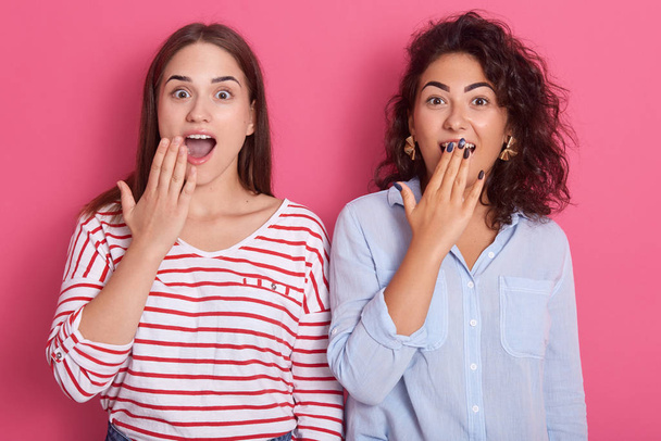 Two surprised cute young women pose together isolated over pink background, beautiful brunette females cover their widely opened mouths, look astonished and surprised, wear casual. Friendship concept. - Foto, immagini