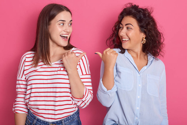 Indoor shot of two young beautiful girls pointing towards each other, brunette women dress shirts, standing against pink studio background, expresses happynes and astonish. Same sex love concept. - Фото, изображение