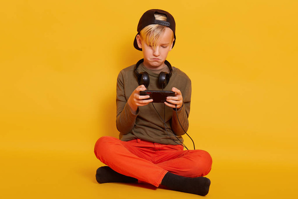 Little boy sitting on floor with smartphone in hands, posing in studio, wearing trousers, cap and jumper, blond male child looks concentrated, playing his favourite online game. Childhood concept. - Фото, изображение