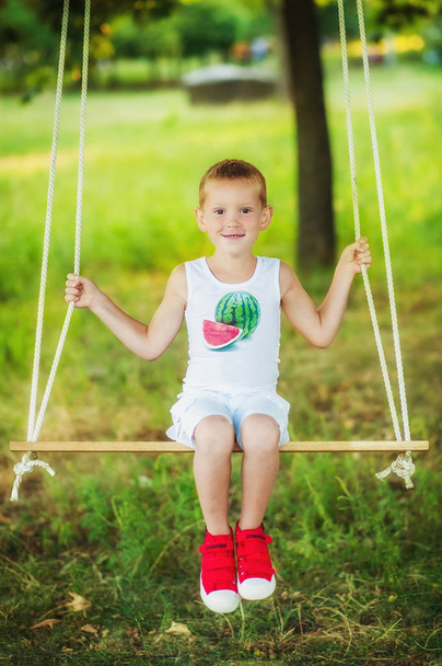 5 years old boy in red sneakers, white shorts and white watermelon printed t-shirt swinging in garden. Swing outdoors for children. Child playing in park in summer time. Kids play - Photo, Image