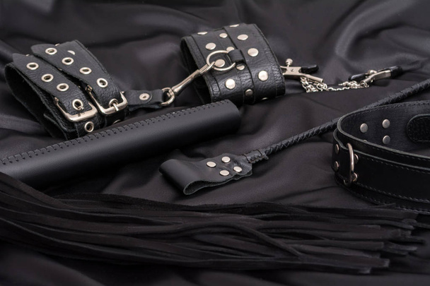 Leather handcuffs, black whip, chain collar and stack on black background. Accessories for adult sexual game. BDSM outfit. - Фото, изображение