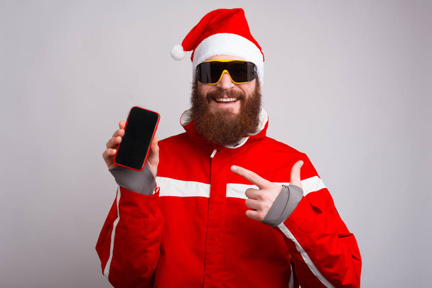 Onnellinen nuori alpinisti yllään joulupukki hattu ja osoittaa älypuhelin
 - Valokuva, kuva