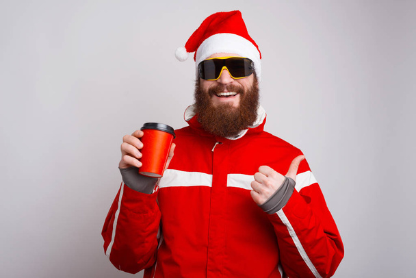Foto de alpinista feliz con sombrero de Santa Claus sosteniendo la taza de café para ir y mostrando los pulgares hacia arriba
 - Foto, Imagen