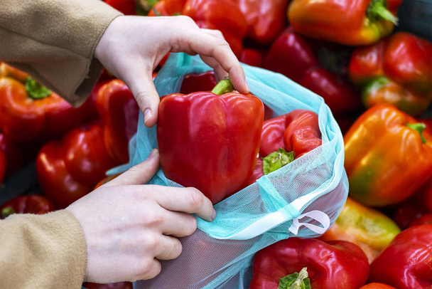 Een vrouw stopt rode paprika 's in een herbruikbare zak uit een grote doos in een groentewinkel. - Foto, afbeelding