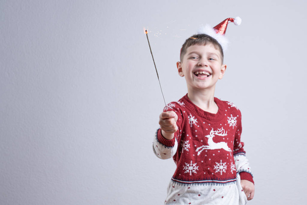 Boldog mosolygós kisfiú piros télapó pulóver és kalap tartja a kezében csillagszóró, elszigetelt szürke háttér, Boldog karácsonyt és boldog új évet koncepció - Fotó, kép