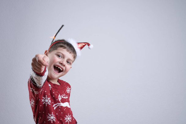 Menino sorridente alegre em suéter vermelho de santa e chapéu segura em sua mão sparkler, isolado sobre fundo cinza, Feliz Natal e conceito de Feliz Ano Novo
 - Foto, Imagem