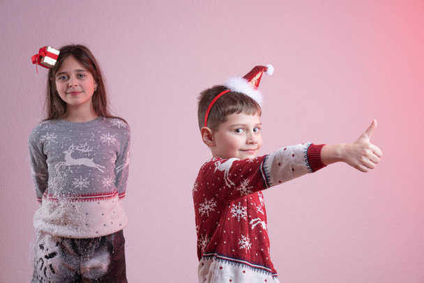 Ευτυχισμένο παιδί, αγόρι με κόκκινο πουλόβερ Σάντα και καπέλο δείχνει σημάδι επιτυχίας και κοιτάζοντας την κάμερα, η αδελφή του με γκρι πουλόβερ Σάντα και καπέλο βαμμένο σε ψεύτικο χιόνι στέκεται στο παρασκήνιο, απομονώνονται πάνω από το ροζ - Φωτογραφία, εικόνα