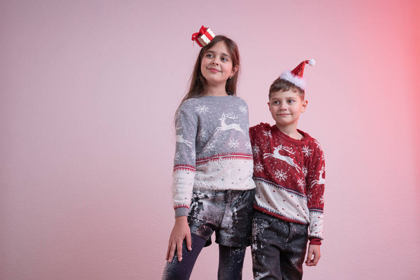 Children in santa sweaters and hats stained in synthetic snow isolated over pink background, brother hugs his sister, Merry Christmas and Happy New Year concept - Fotó, kép
