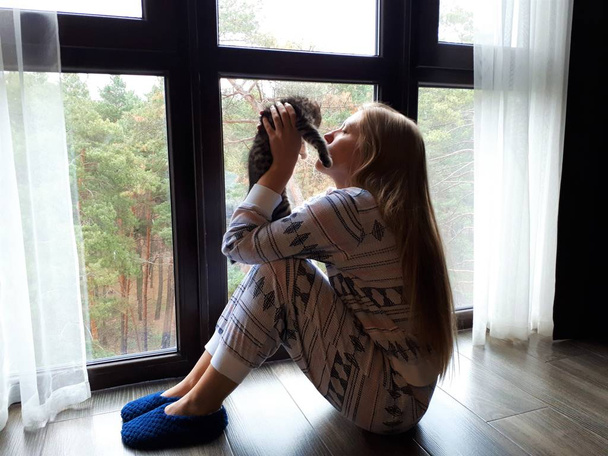 mooi meisje met blond lang haar in pyjama zit bij het raam met een grijze beker en een katje - Foto, afbeelding