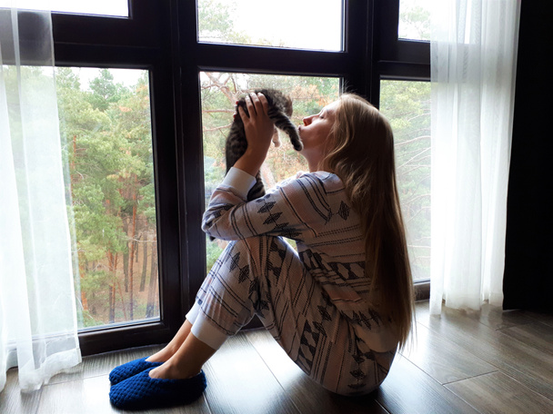 hellhäutiges junges Mädchen mit blonden langen Haaren im Schlafanzug sitzt am Fenster mit einer grauen Tasse und einem Kätzchen - Foto, Bild