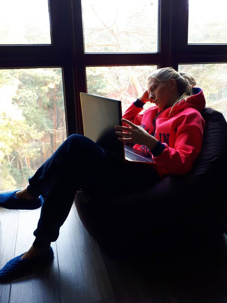 молода блондинка вдома працює на ноутбуці і розмовляє по телефону біля великого вікна
 - Фото, зображення