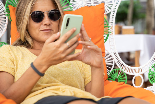Menina loira com óculos de sol deitada em um peito de bambu olhando para um celular - Foto, Imagem