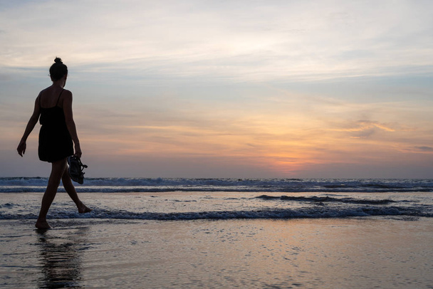 Девушка, идущая по воде на пляже с ботинками в руке, когда солнце садится - Фото, изображение