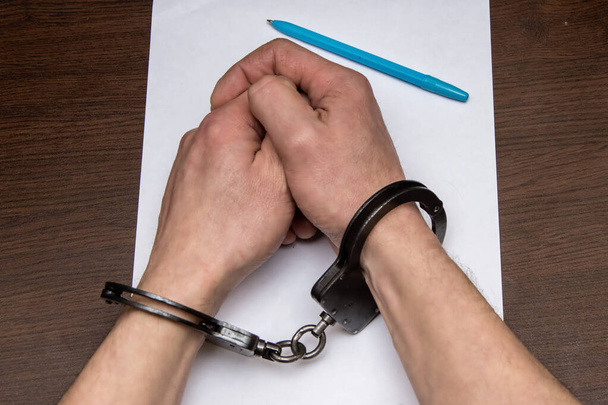 Ένας άντρας με γυμνά χέρια με χειροπέδες κάθεται σε ένα τραπέζι μπροστά από ένα λευκό φύλλο χαρτί και ένα στυλό. Έννοια: ο κρατούμενος γράφει μια ομολογία, ο κρατούμενος υπογράφει το έγγραφο. - Φωτογραφία, εικόνα