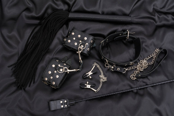 Leather handcuffs, black whip, chain collar, nipple clamps and stack on black background. - Foto, Imagem