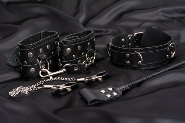 Leather handcuffs, nipple clamps, chain collar and stack on black silk background. Accessories for adult sexual game. BDSM outfit. - Fotó, kép