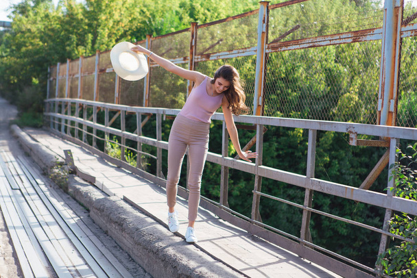 Pretty young woman wearing cowboy hat posing on the old rusty bridge during sunset. - 写真・画像