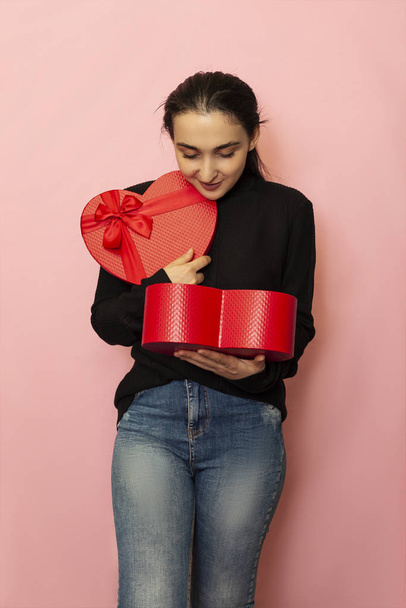 Emocje twarzy. Niesamowita, zaskoczona kobieta trzyma pudełko prezentów w kształcie serca. Atrakcyjny model mody w odzieży casual pozowanie z czerwonym pudełku prezentu. Prezent na Walentynki. Dzień miłości, rocznica - Zdjęcie, obraz