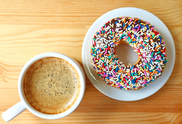 Top view of a cup of hot coffee and a donut topped with colorful sprinkles served on wooden table - Valokuva, kuva