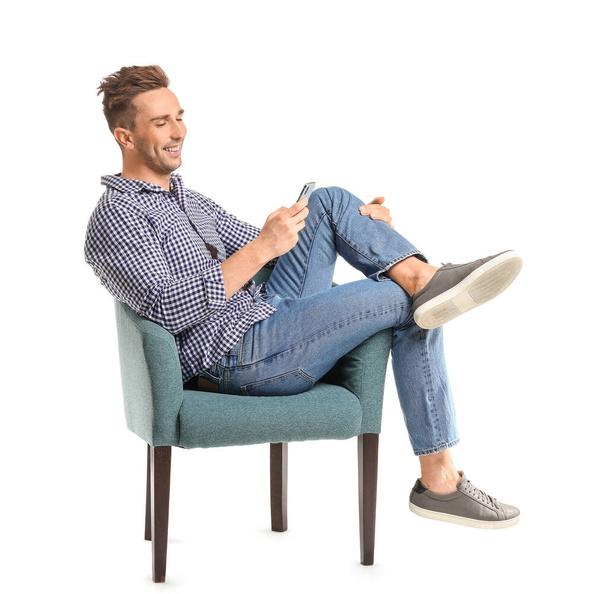 Knappe man met mobiele telefoon zittend in fauteuil op witte achtergrond - Foto, afbeelding