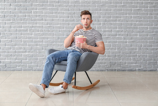Шокированный человек смотрит кино, сидя в кресле возле серой стены
 - Фото, изображение