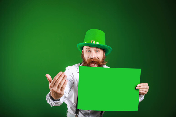 Bearded man with blank poster inviting viewer on color background. St. Patrick's Day celebration - Fotografie, Obrázek