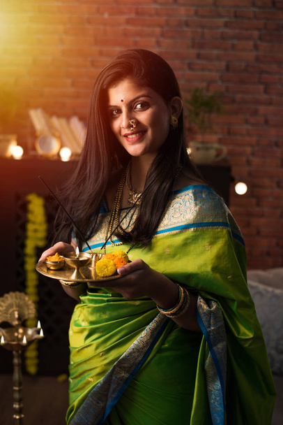 Indian Girl / women holding Pooja Thali while wearing green saree and sitting at home with flower rangoli and Samai, celebrating Diwali or hindu festival. selective focus - Photo, image