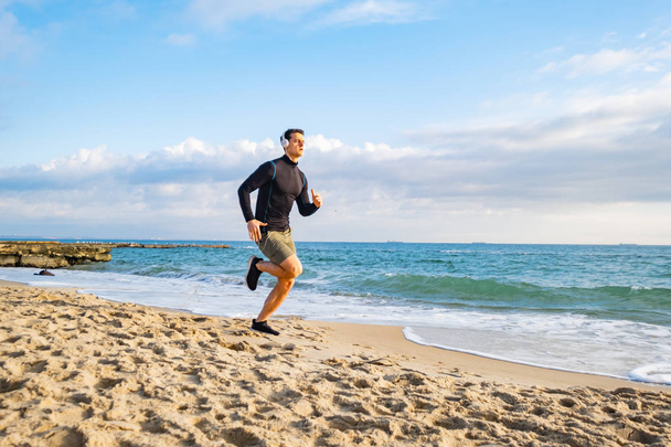 Fit male runner training on the summer beach and listen to music against beautidul sky and sea - Foto, Imagen