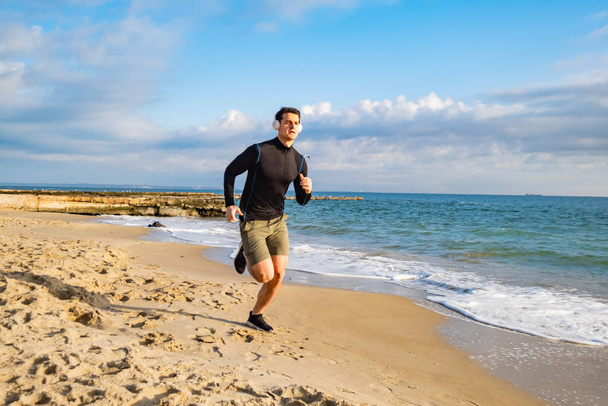 Fit male runner training on the summer beach and listen to music against beautidul sky and sea - Фото, изображение