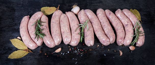 Неприготовленные свиные колбаски среди специй на темной поверхности, вид сверху
 - Фото, изображение
